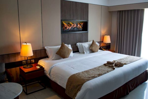 A well-decorated hotel room features a large bed with brown accents, bedside tables with lamps, and a wall-mounted fireplace.