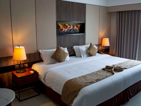 A well-decorated hotel room features a large bed with brown accents, bedside tables with lamps, and a wall-mounted fireplace.