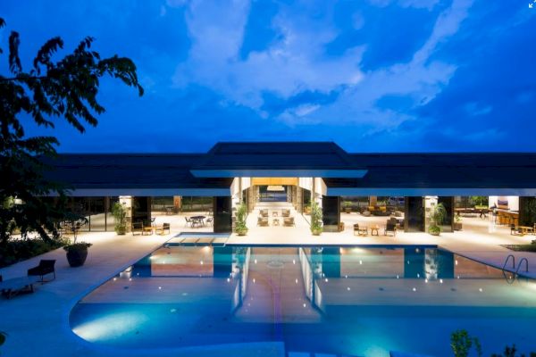 A luxurious, modern building with a large swimming pool at twilight, featuring extensive lighting and an open-air design.