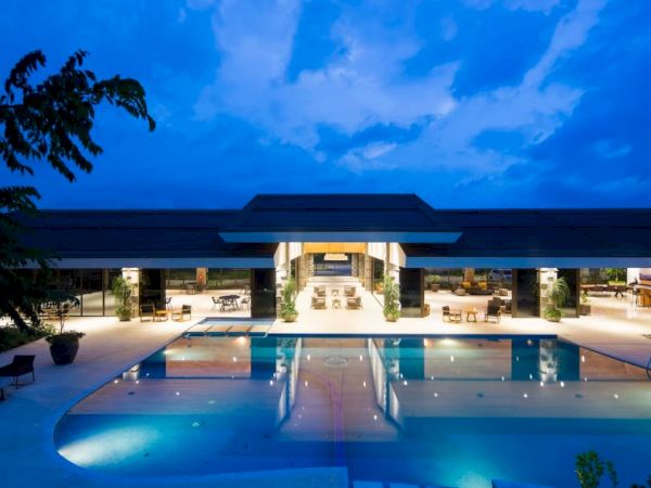 A luxurious, modern building with a large swimming pool at twilight, featuring extensive lighting and an open-air design.