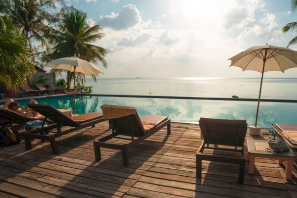 The image shows loungers and umbrellas on a wooden deck beside an infinity pool with a scenic ocean view and palm trees in the background.