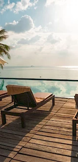 A serene poolside scene with lounge chairs, umbrellas, and a calm infinity pool overlooking an ocean view, with palm trees and a sunny sky.
