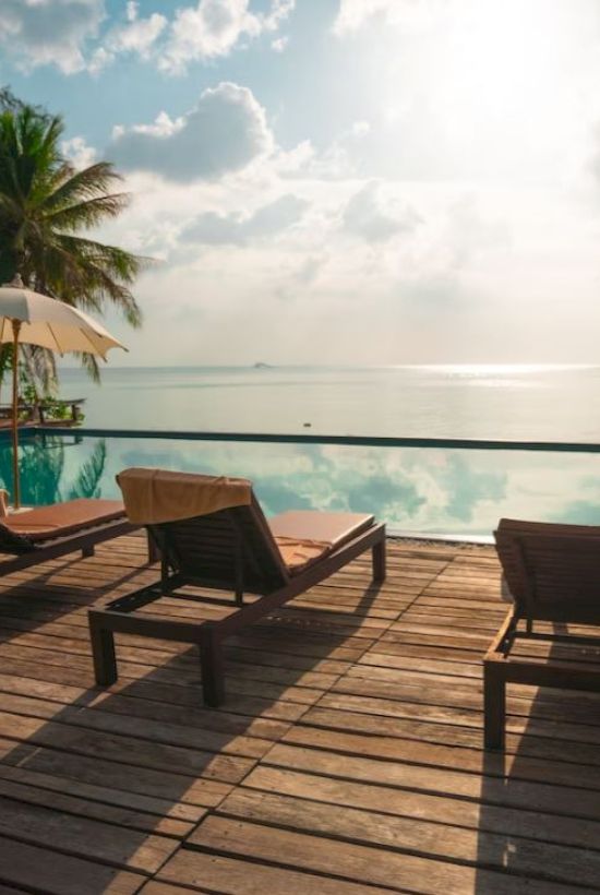 A serene poolside scene features lounge chairs, umbrellas, palm trees, and a stunning view of the ocean with a clear, sunny sky in the background.