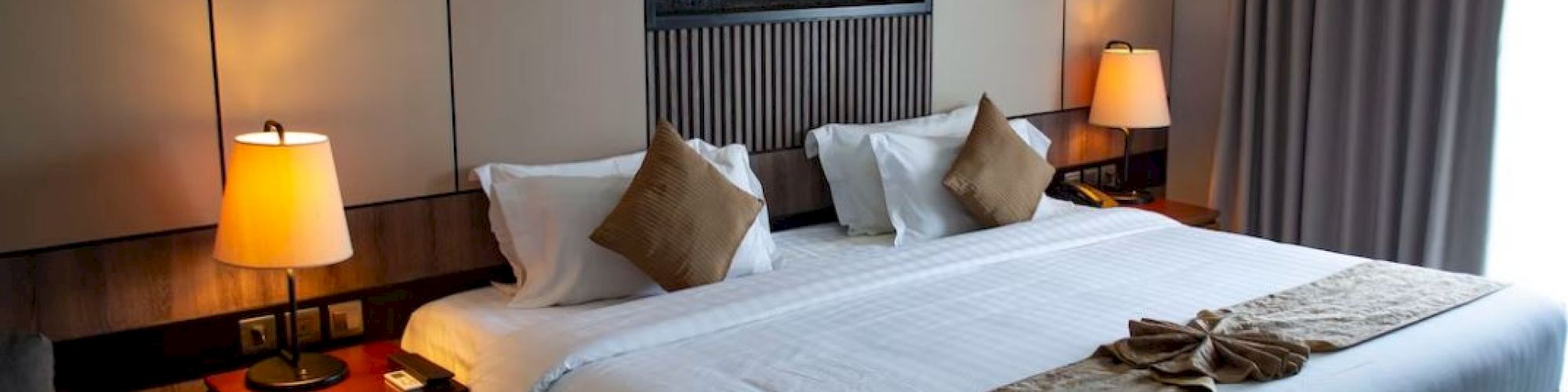A modern hotel room with a large bed, two bedside lamps, decorative pillows, and a painting above the bed. Curtains cover the window.