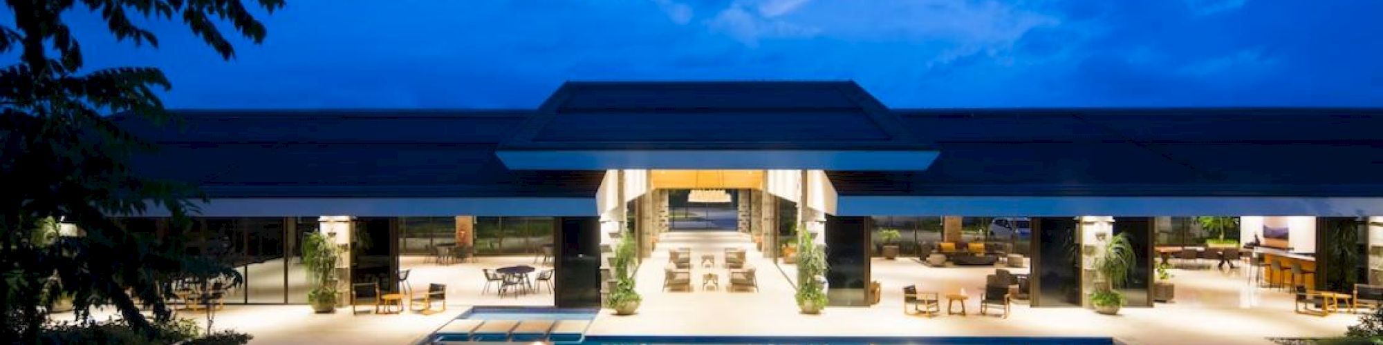 The image shows a nighttime view of a modern luxury building with an illuminated swimming pool and surrounding lounge area, set against a blue sky.