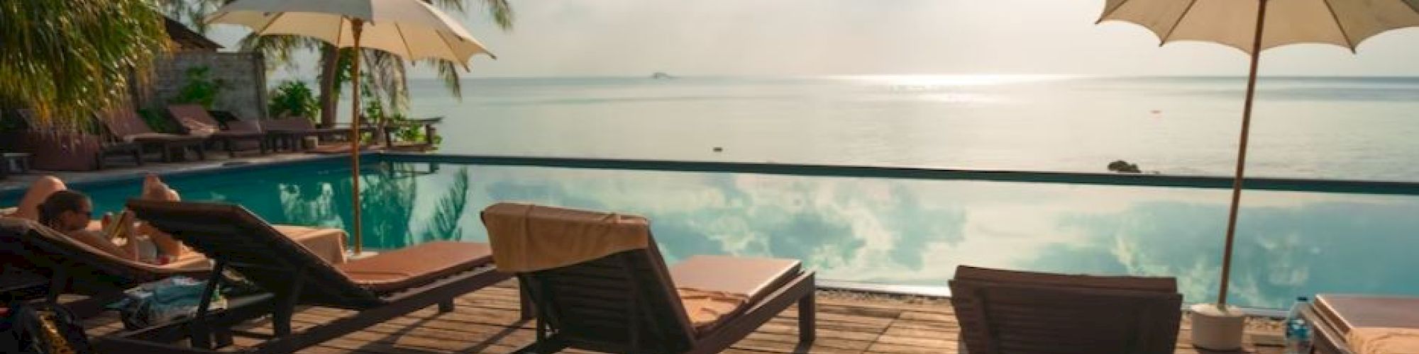 A serene tropical scene showing lounge chairs and umbrellas by a pool, with palm trees and the ocean in the background, under a bright sky.