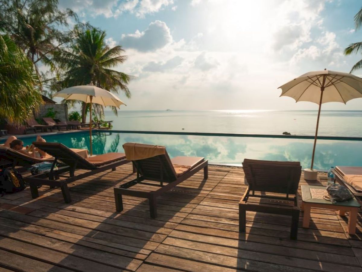 Poolside deck with lounge chairs and umbrellas overlooking a serene body of water, surrounded by palm trees and clear skies.