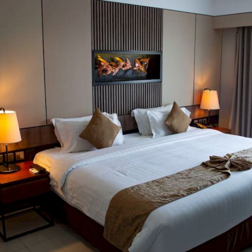 This image shows a well-lit, contemporary hotel room with a large bed, bedside tables, lamps, and a decorative wall art above the headboard.