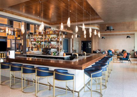 This image shows a modern bar with high blue chairs, a well-stocked back bar, pendant lighting, and a spacious, stylish seating area.