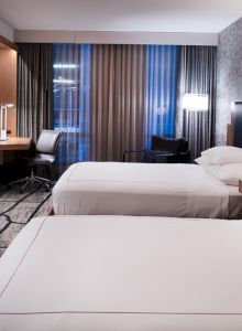 A modern hotel room with two double beds, a flat-screen TV, a desk with a chair, nightstands, and contemporary decor, featuring abstract patterns.