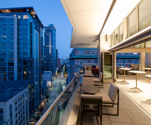 A modern balcony overlooking a city street with tall buildings, furnished with tables and chairs, under a clear evening sky.