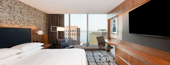 A modern hotel room with a large bed, a desk with a chair, a flat-screen TV, and a view of buildings outside through a large window.