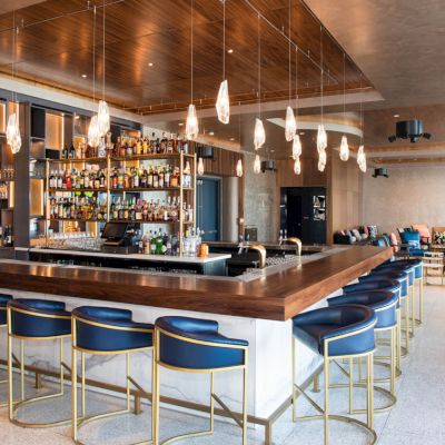 A modern bar with a well-stocked liquor shelf, blue bar stools with gold accents, and stylish pendant lights. Comfortable seating in the background.