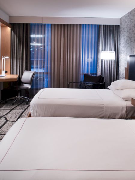 The image shows a modern hotel room with two neatly made beds, a flat-screen TV, a desk with a chair, and a well-lit sitting area by the window.