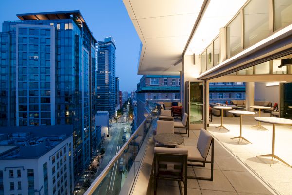 A modern cityscape view from a high-rise balcony at dusk, featuring seating and tables, glass railings, and adjacent tall buildings.