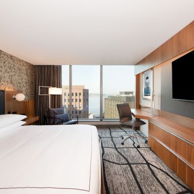 A modern hotel room with a large bed, a desk, chair, and a wall-mounted TV. The window offers a city view with buildings in the background.