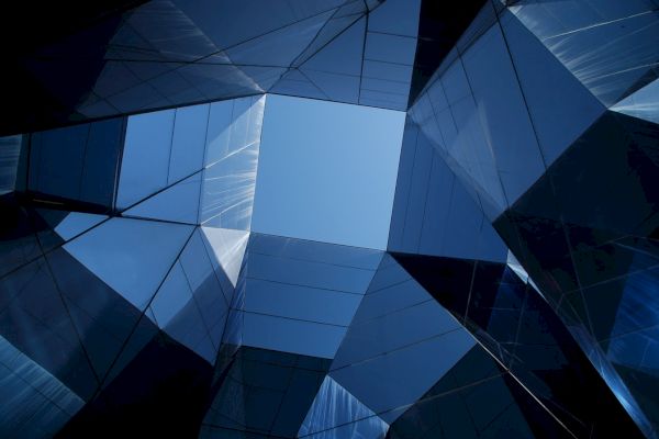 The image shows a geometric architectural structure with glass panels reflecting the sky, creating a mosaic-like view looking upwards.