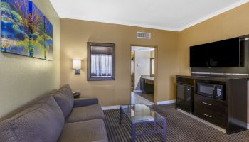 A cozy living room with a gray sofa, glass coffee table, flat-screen TV, colorful wall art, lamp, and a small kitchenette area.