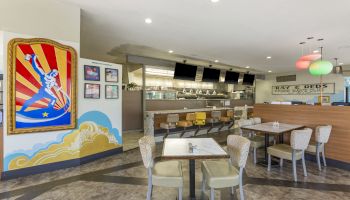 A bright diner with colorful wall art, tables and chairs, a counter with stools, and hanging lights. The seating area is cozy and inviting.