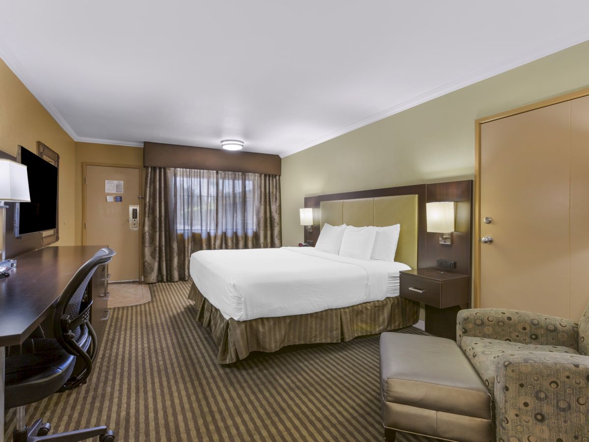 A hotel room with a double bed, desk, chair, lamp, TV, and an armchair with an ottoman, featuring beige tones and carpeted floor.