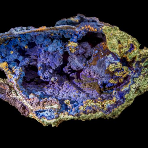 The image shows a vibrant, multicolored mineral specimen with a mix of purple, blue, green, and yellow hues against a black background.