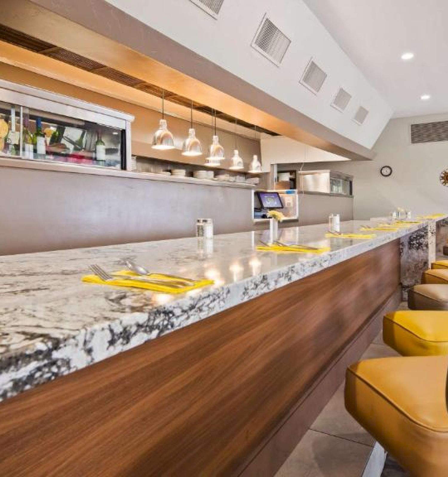 This image shows a modern restaurant interior with a marbled counter, yellow chairs, hanging lights, and framed art on the wall in the background.