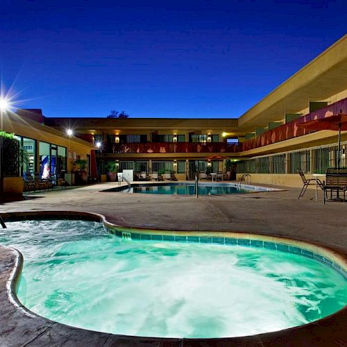 Exterior night view of a hotel featuring a lit jacuzzi and pool, surrounded by a courtyard with tables, chairs, and a two-story building.