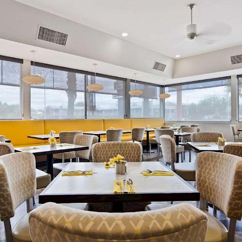 A modern restaurant with yellow and beige decor, featuring tables with yellow napkins, flowers, and chairs arranged in a clean, well-lit dining space.