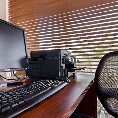 An office workspace with a Dell monitor, keyboard, printer, and a mesh chair, set on a wooden desk with window blinds in the background.