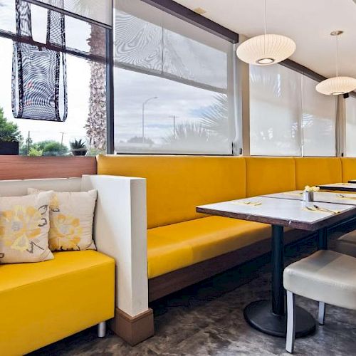 An empty restaurant features a long yellow booth with floral cushions, modern hanging lamps, and tables set with silverware.