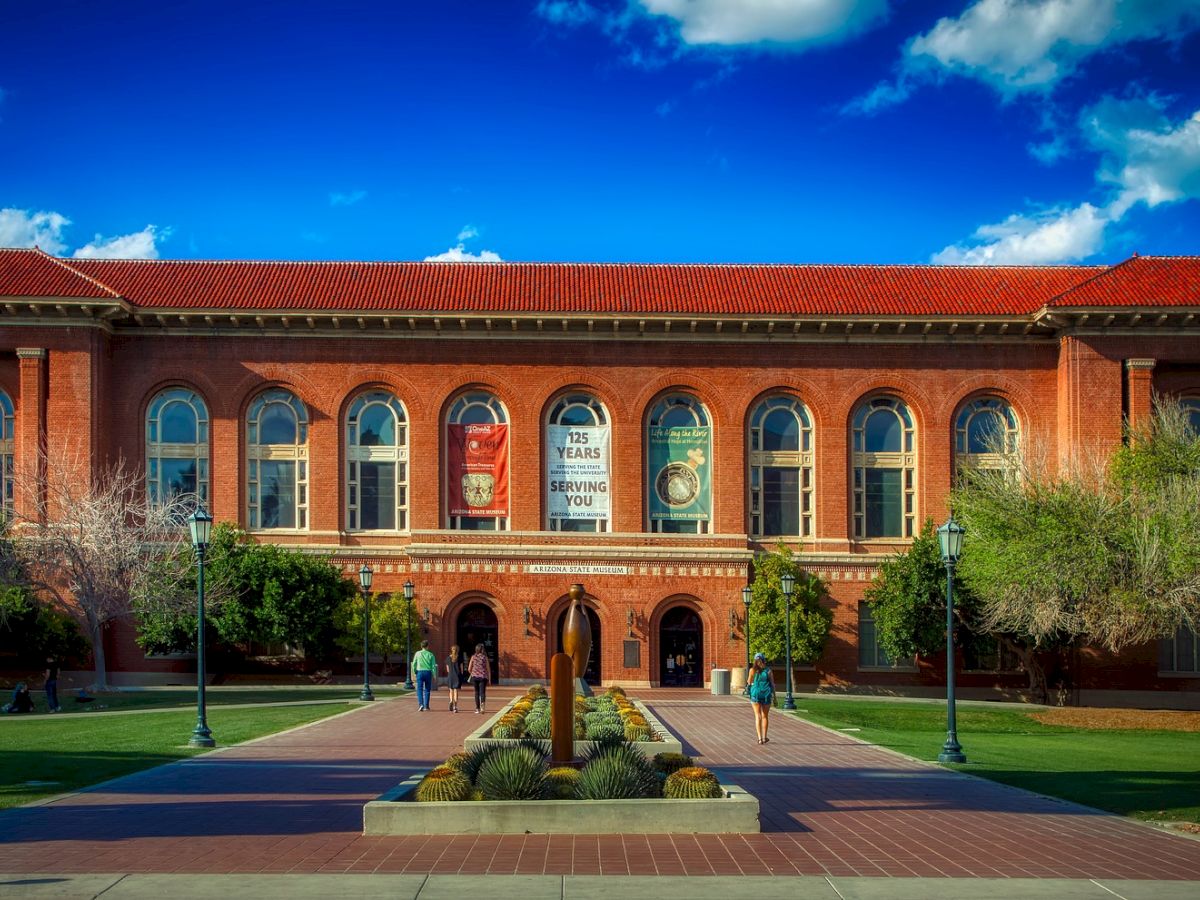 A red brick building with arched windows, a landscaped walkway, and a fountain in front, with people walking and blue sky above.