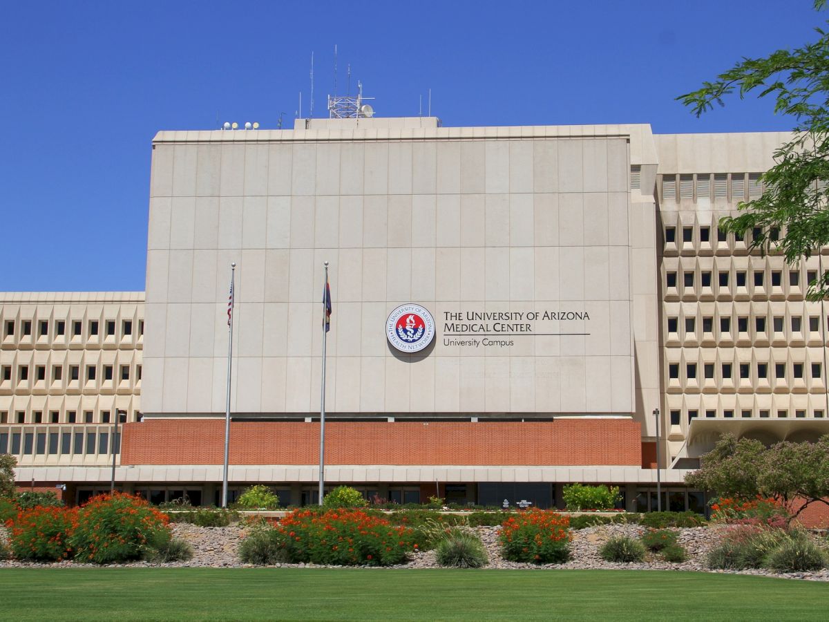 The image shows the University of Arizona Medical Center, a large beige building with multiple windows and a landscaped garden in the foreground.