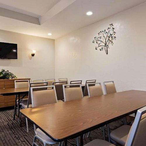 A meeting room features a long table with chairs, a wall-mounted TV, a cabinet, plants, and a decorative tree wall art.