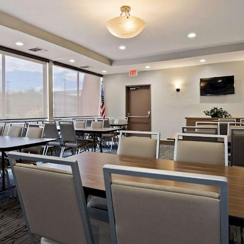 A conference room with multiple tables and chairs, a wall-mounted TV, and large windows with blinds.