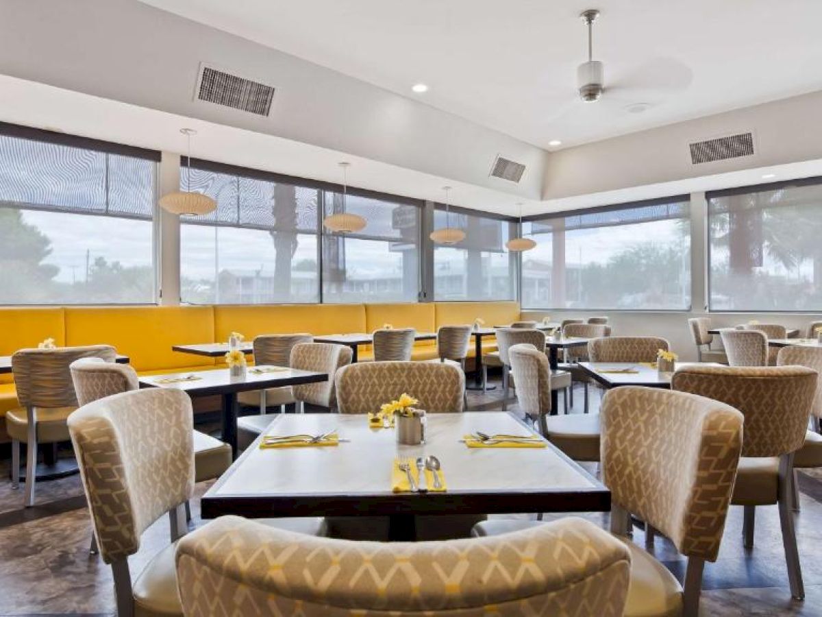 A modern dining area with yellow booths, cushioned chairs, and neatly set tables with yellow napkins and small vases of flowers, and large windows.