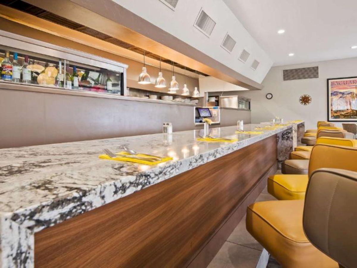 A modern bar or restaurant interior with a marble countertop, yellow chairs, hanging lights, a liquor shelf, and a poster on the wall is visible.