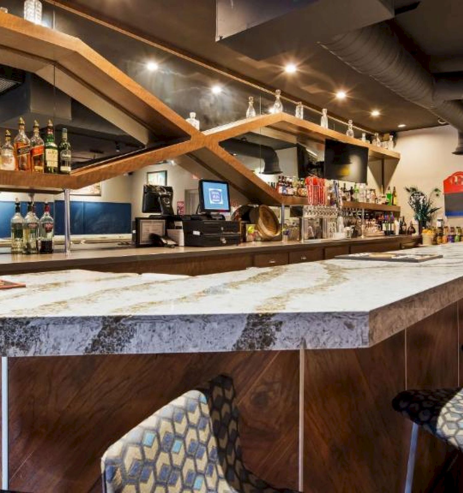 The image shows a modern bar with a long counter, a variety of alcohol bottles, barstools, and decorative elements on the walls.