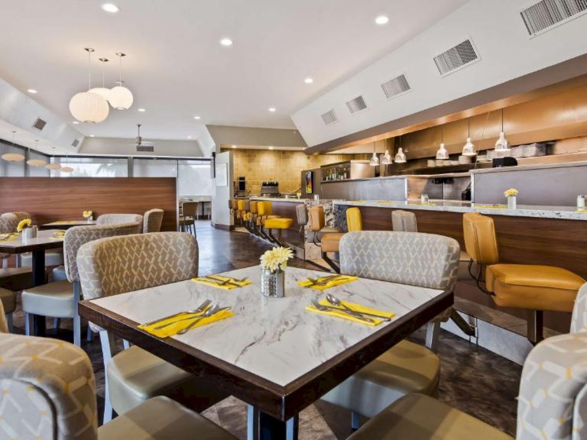 The image shows a modern, well-lit restaurant interior with patterned chairs, marble-topped tables, and yellow accents, giving a bright, inviting feel.