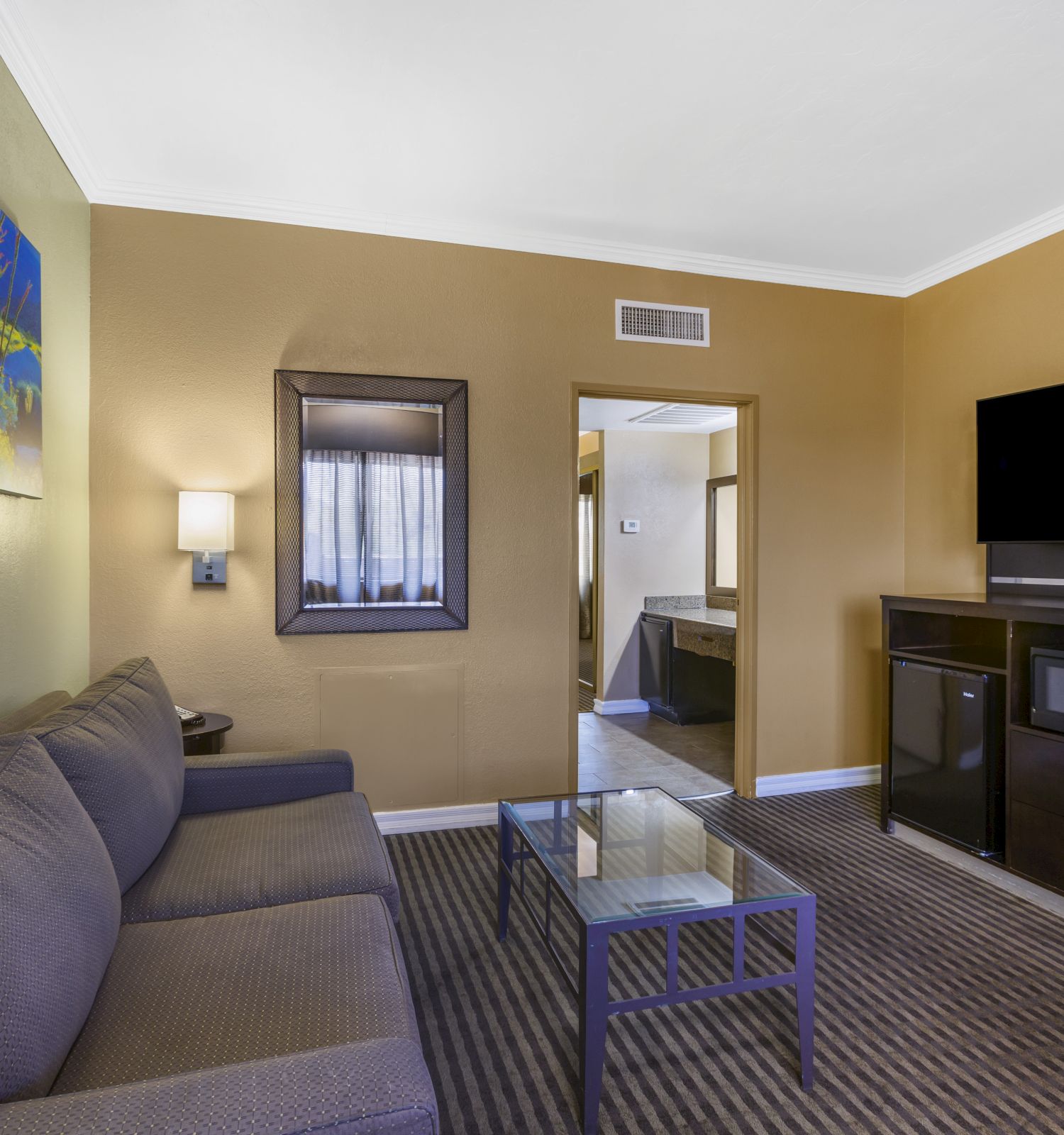 A living room with a sofa, glass coffee table, wall art, TV, microwave, fridge, and neutral colors, leading into another room.