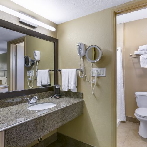 A bathroom with a large mirror, sink, hairdryer, towels, and a separate area containing a toilet and shower with towels on a shelf above.