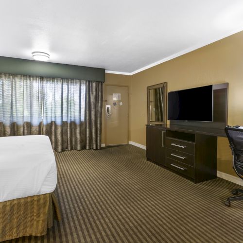 A hotel room with a bed, TV on a console, an office chair, desk, lamp, mirror, and curtains covering the window, ending the sentence.