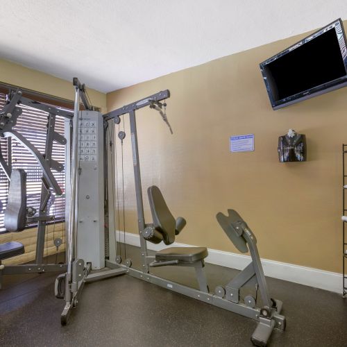 The image shows a gym with a multi-station workout machine, TV on the wall, shelves with towels, and a window with blinds.