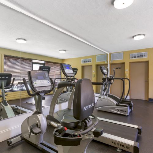 The image shows a gym with exercise bikes, treadmills, a wall mirror, and a water dispenser in a well-lit room.