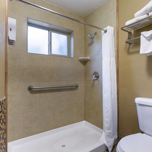 A bathroom with a walk-in shower, grab bar, white curtain, window, toilet, towel rack with folded towels, and beige walls decorated with tiles.