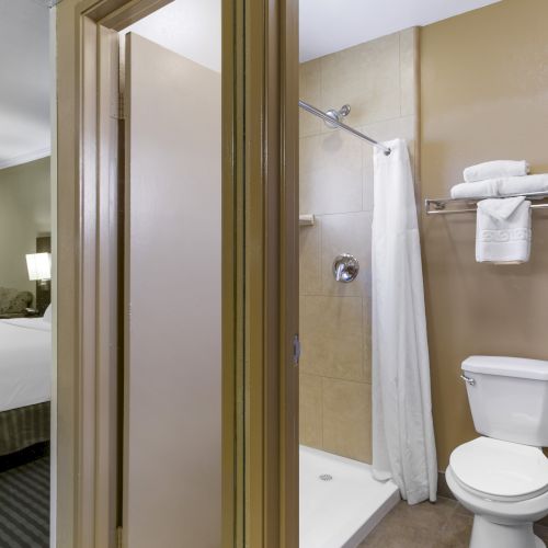 The image shows an interior view of a hotel room, focusing on the bathroom with a toilet, shower, and door, adjacent to the bedroom area.