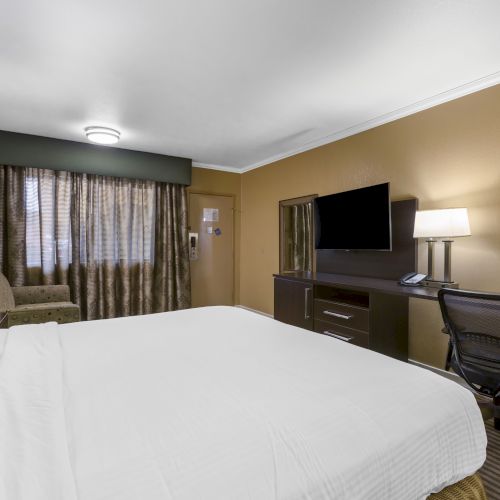 A hotel room with a bed, desk, chair, TV, couch, lamps, and curtains, decorated in neutral tones. The room appears clean and well-lit.