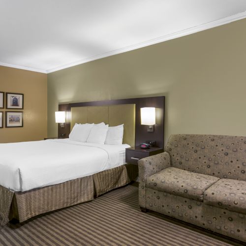 A hotel room with a bed, bedside tables, lamps, framed pictures on the wall, and a beige sofa on a striped carpet floor is shown.