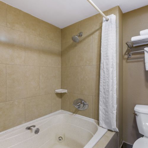 The image shows a bathroom with a bathtub, showerhead, shower curtain, toilet, and neatly folded towels on a wall-mounted rack.