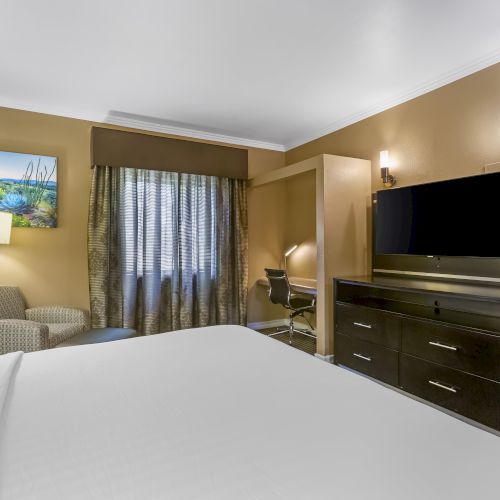 A well-lit hotel room with a bed, TV, desk, chair, and a window with curtains. There are lamps and a painting on the wall.