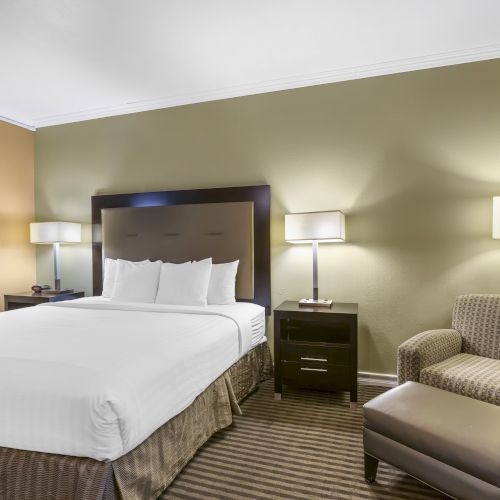 A neatly furnished hotel room with a bed, nightstands, lamps, armchair, ottoman, and wall art, all in a warm, inviting color scheme.